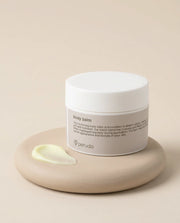 A jar of Peruda body balm with a white lid and beige label, resting on a matching beige dish with a dollop of pale yellow balm in front, set against a solid beige background.