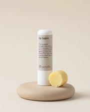 A white Peruda lip balm tube stands upright on a round beige stone base next to a round yellow balm piece, against a warm beige background.