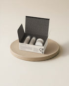 An open white and black patterned box containing three small cosmetic tubes, displayed on a round beige ceramic plate against a warm off-white background.