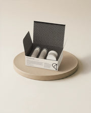 An open white and black patterned box containing three small cosmetic tubes, displayed on a round beige ceramic plate against a warm off-white background.