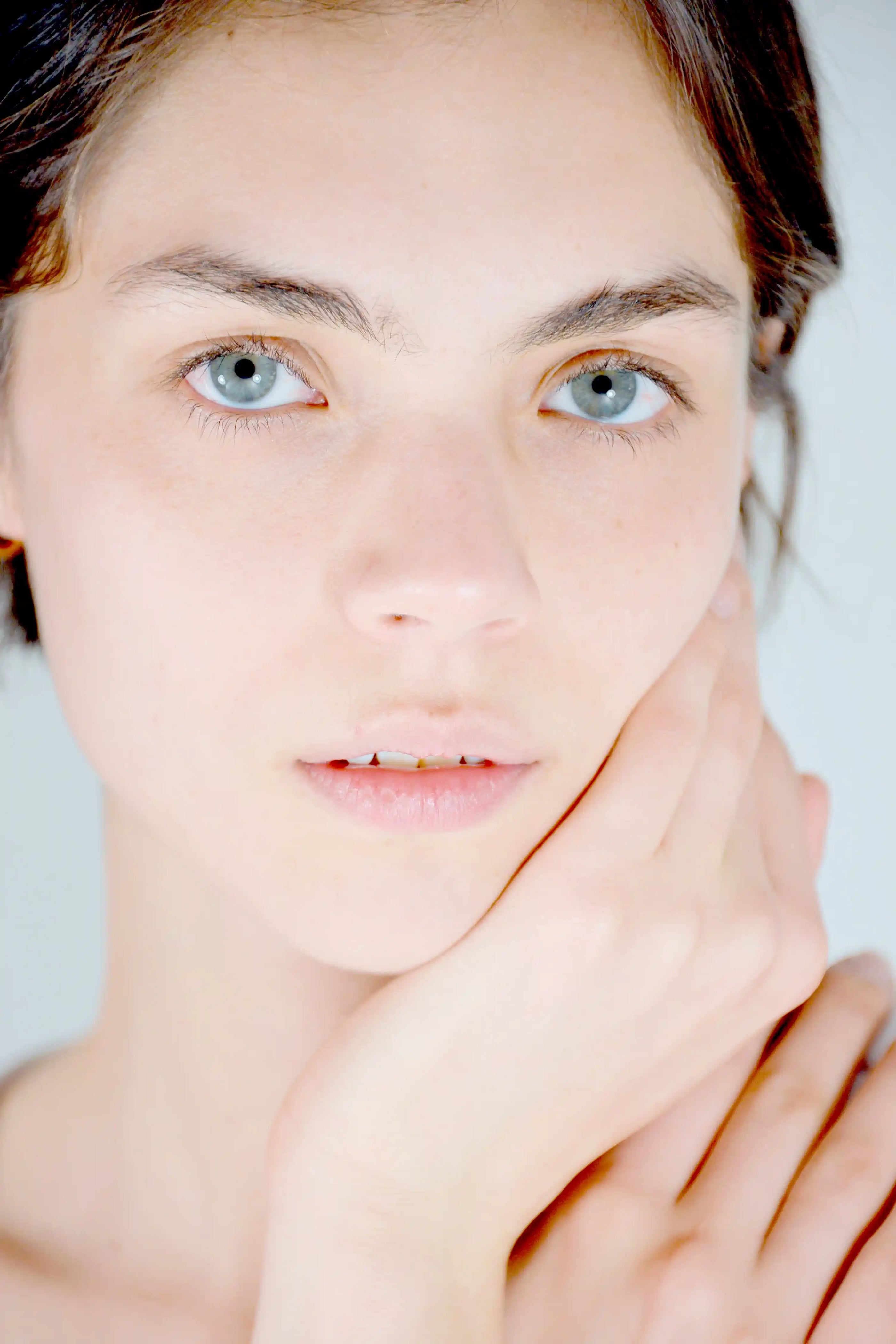 Close-up portrait of a woman with light blue eyes and fair skin, touching her cheek with one hand against a soft light gray background.