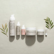 Six skincare products from the brand Typhosa are arranged in a row on a textured light beige fabric background, flanked by decorative olive branches. The products include a hair wash, neck oil, face wash, body wash, body butter, and a small tube, all in neutral white and pale pink packaging with black text.