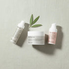 Three Peruda skincare products arranged on a textured light beige surface: a face oil bottle, a healing mask jar, and a face serum bottle, with a green leaf sprig between them.