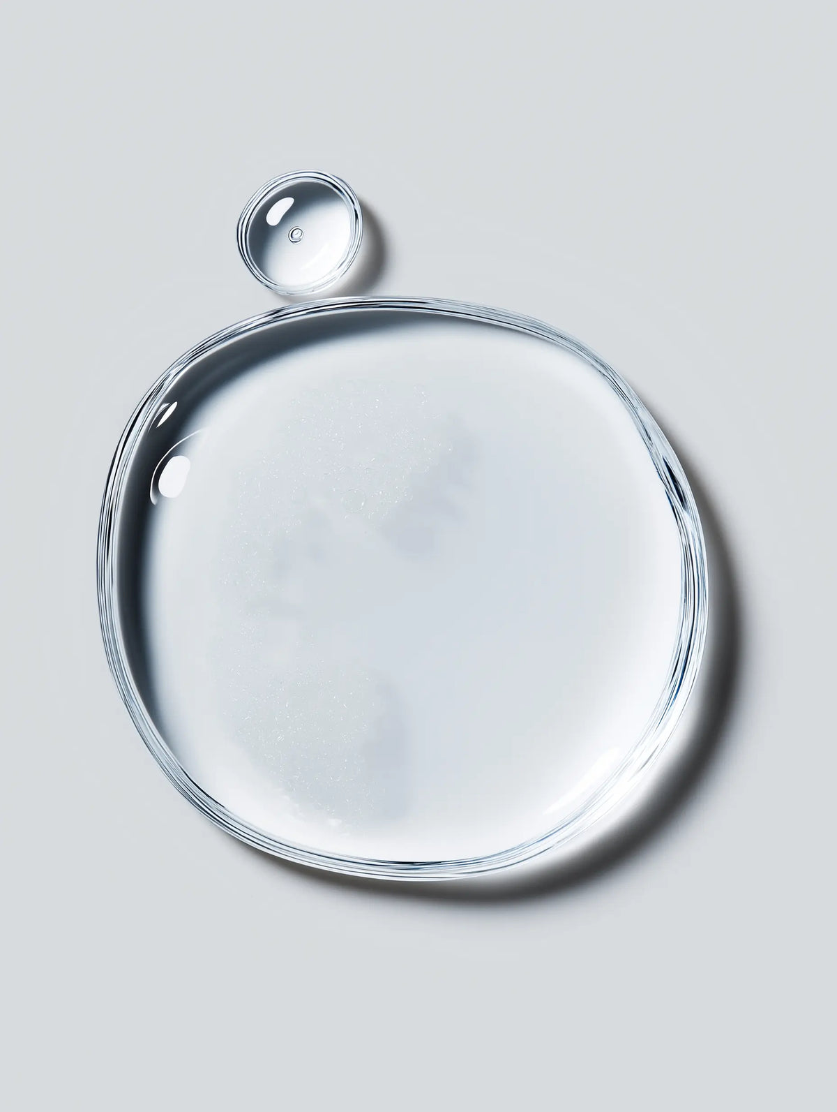 A clear, transparent gel or liquid droplet with a glossy finish, arranged in a large irregular circle with a smaller bubble above it, set against a solid light gray background.