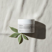 peruda skincare body balm jar with water droplets on a textured white fabric background with green leaves